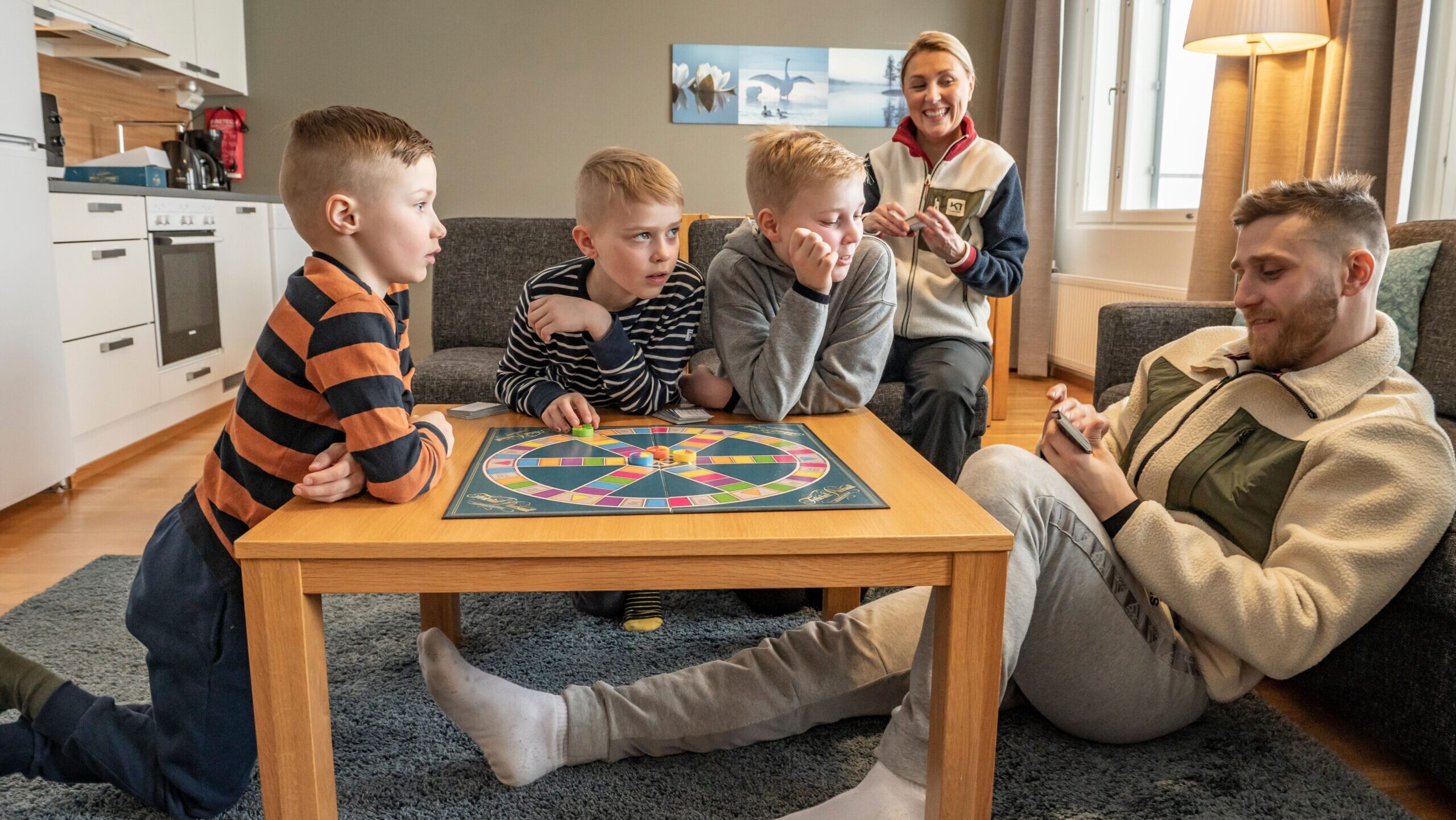 Perhe istuu pöydän ääressä pelaamassa Trivial Pursuit -lautapeliä.